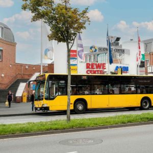 Vor dem Haupteingang des Seidnitz Centers hält ein gelber Linienbus. Im Hintergrund sind die bunten Flaggen vor dem Center zu sehen.