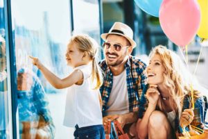 Eine junge Familie mit Mutter, Vater und etwa 4-jähriger Tochter schaut begeistert in das Schaufenster eines Shoppingcenters. Die Mutter hält bunte Ballons an Schnüren.