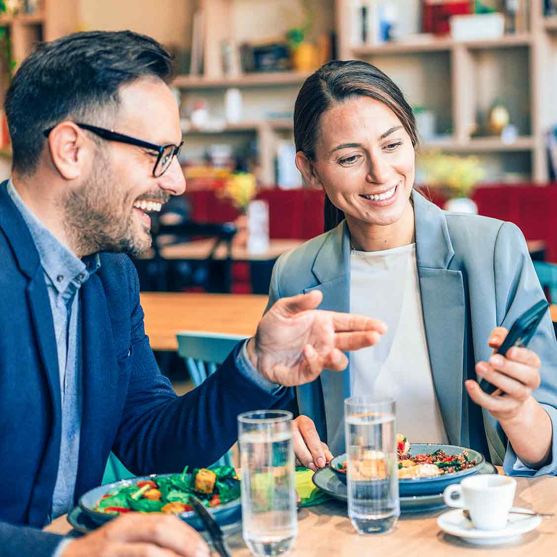 Eine Frau und ein Mann in legerer Business-Kleidung sitzen an einem Tisch und schauen lachend gemeinsam auf ein Smartphone. Auf em Tisch stehen Speisen, Gläser mit Waser und eine Tasse Espresso.