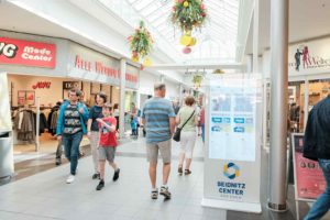 Mehrere Personen laufen durch einen Gang des Seidnitz Centers. Im Vordergrund steht eine Stele mit dem Centerplan. Von der Decke hängt Dekoration mit übergroßen künstlichen Äpfeln.