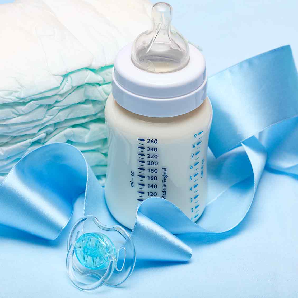 Eine Milchflasche für Babys steht auf blauem Untergrund. Daneben liegen Wegwerfwindeln, ein Schnuller und ein blaues Seidenband.