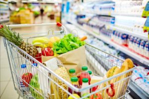 In einem Supermarkt steht ein Einkaufswagen vor einem Regal mit Molkereiprodukten. Der Wagen ist reichlich gefüllt, unter anderem mit Obst, Gemüse, Säften und Brot.