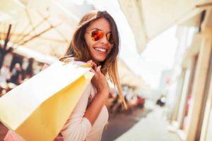 Eine junge Frau mit Sonnenbrille und langen brauen Haaren lächelt in die Kamera. Sie trägt zwei Einkaufstüten aus Papier lässig über der Schulter.